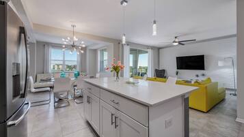 A modern kitchen with a central island, dining area with a sofa, and a living room with a TV and yellow sofa.