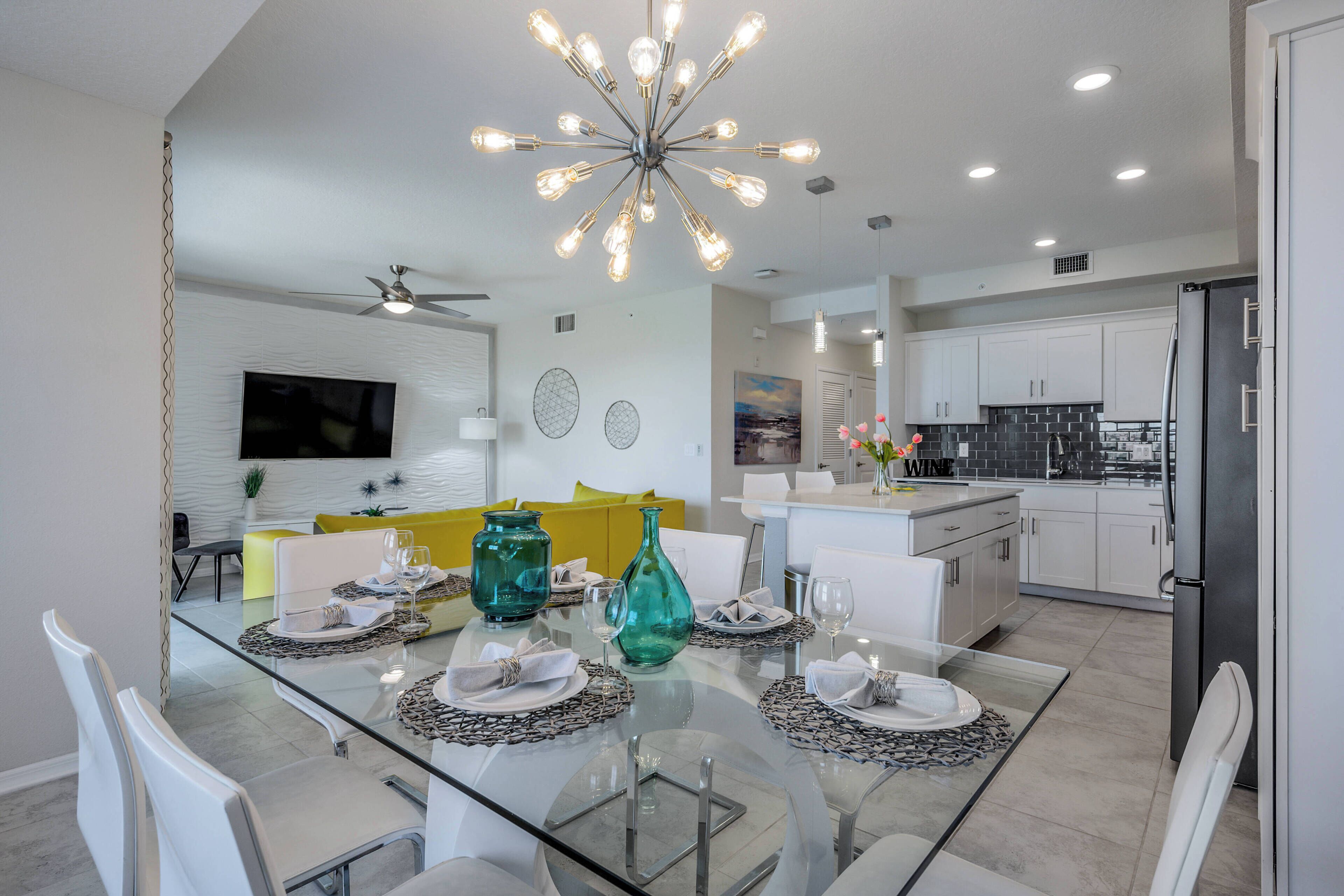 Una cocina moderna con armarios blancos, un refrigerador negro y un comedor con mesa de vidrio y sillas blancas.