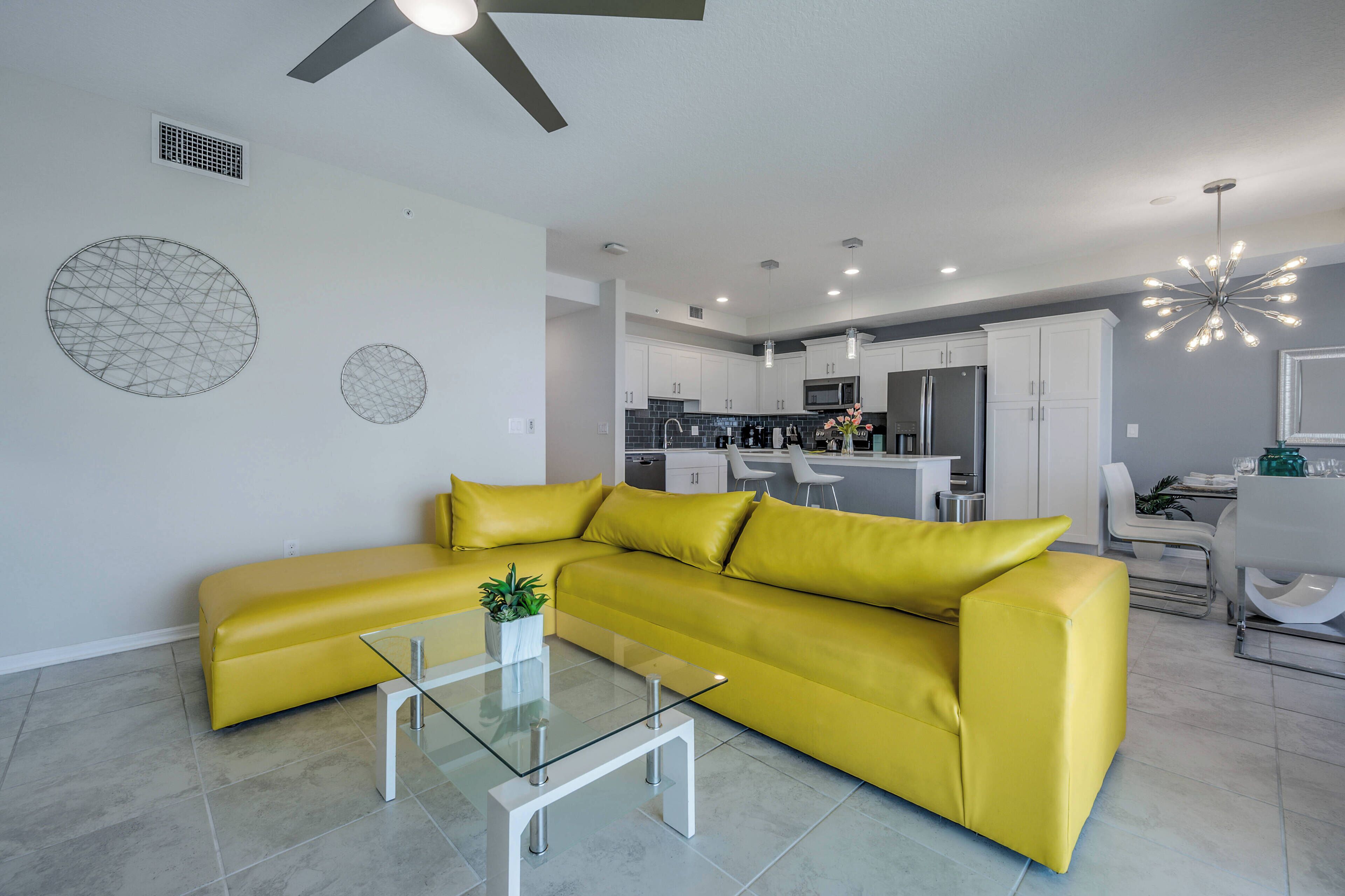 Una sala de estar moderna con un sofá seccional amarillo, una mesa de centro de vidrio y una cocina al fondo.