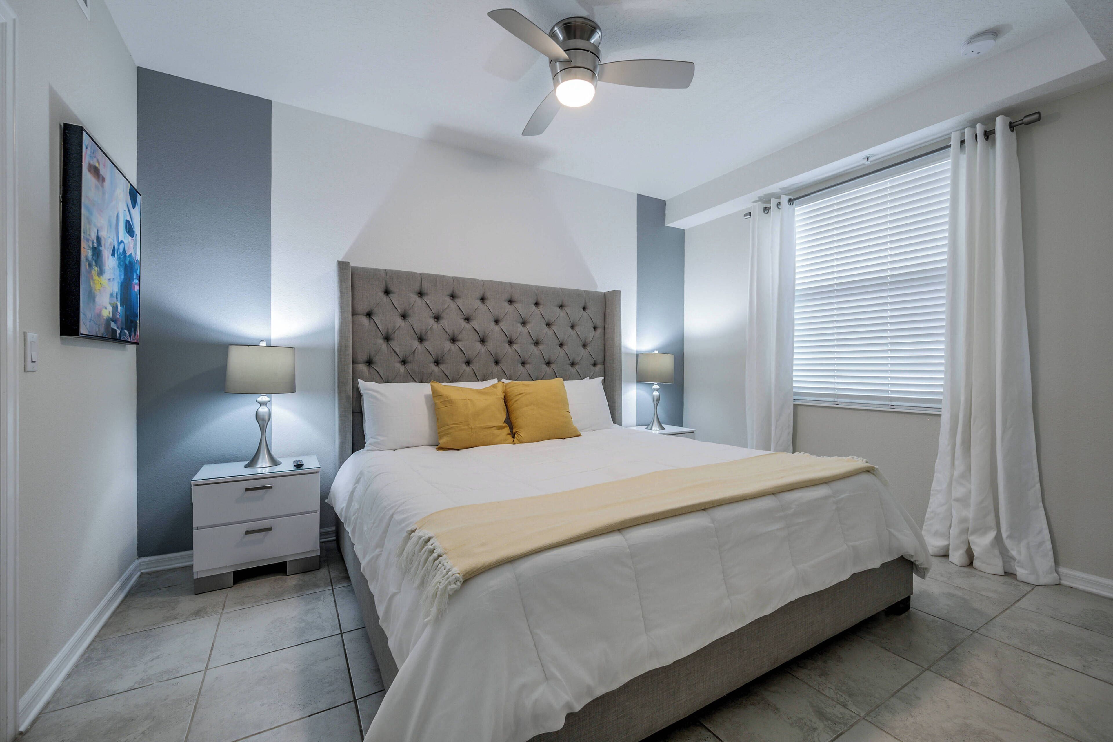 Un dormitorio con cabecera capitonada, cama con ropa de cama blanca y amarilla, dos mesitas de noche con lámparas y un ventilador de techo.