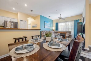 A dining area with a wooden table set for four, a kitchen in the background, and a ceiling fan. - Ow2819 - 2BR Apartment With Balcony Near Disney (Kissimmee)
