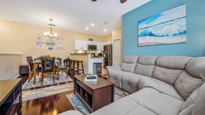 A modern living room with a beige sofa, wooden dining table, and chairs. There is a kitchen area with white cabinets and a microwave. A painting of a beach scene is on the wall. - Ow2819 - 2BR Apartment With Balcony Near Disney (Kissimmee)