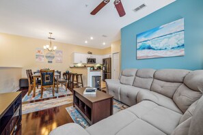 A modern living room with a beige sofa, wooden dining table, and chairs. There is a kitchen area with white cabinets and a microwave. A painting of a beach scene is on the wall. - Ow2819 - 2BR Apartment With Balcony Near Disney (Kissimmee)