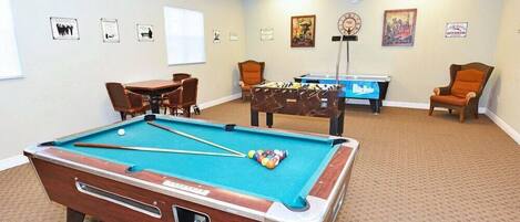A pool table with a pool cue and balls, a smaller pool table, and two chairs.