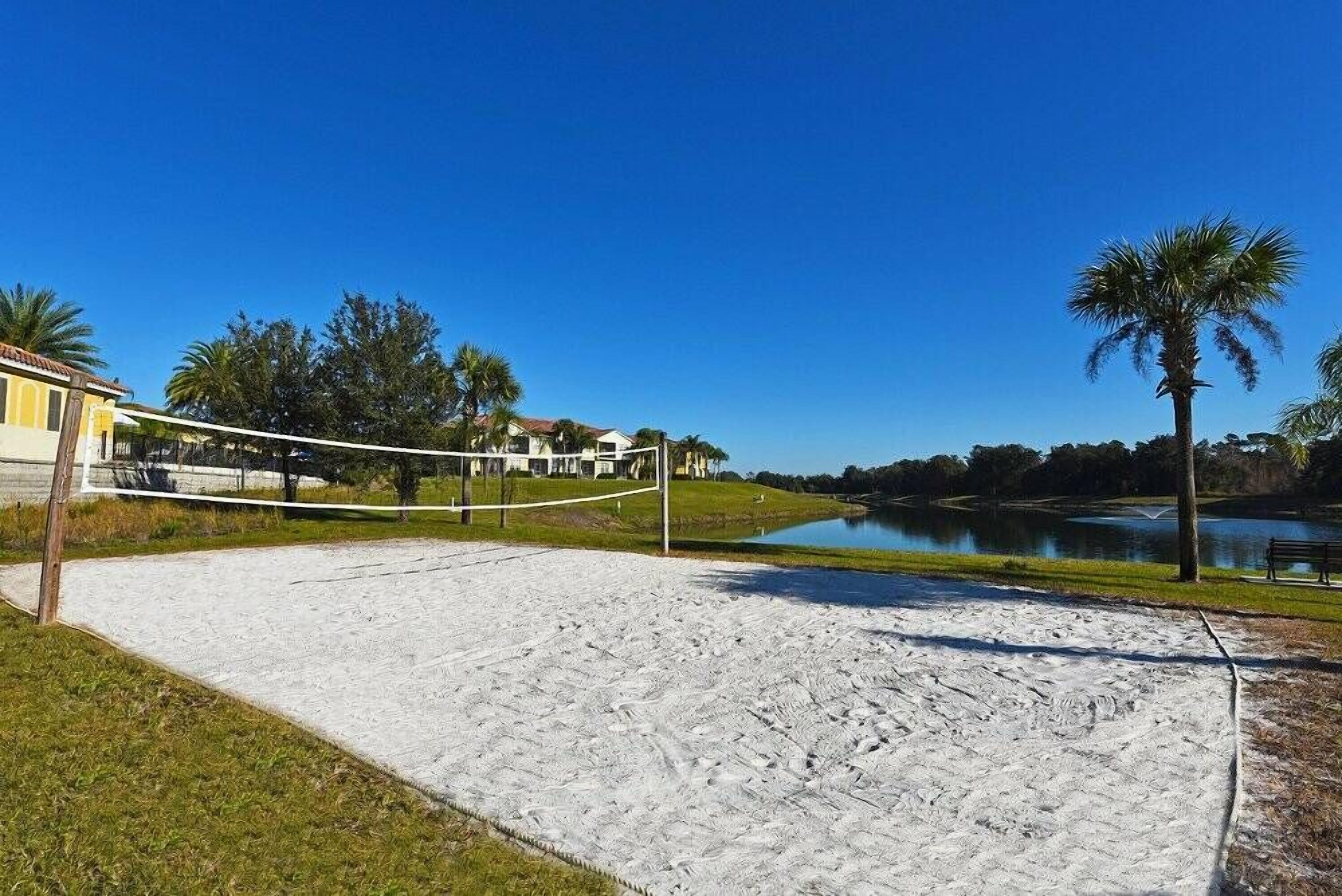 Cancha de voleibol de arena con red, rodeada por un lago, palmeras y un edificio al fondo.