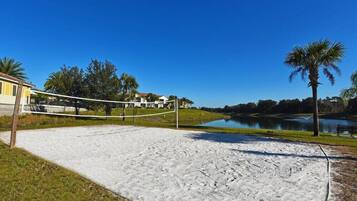 Cancha de voleibol de arena con red, rodeada por un lago, palmeras y un edificio al fondo.
