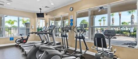 A gym with various cardio machines, large windows, and a ceiling fan.