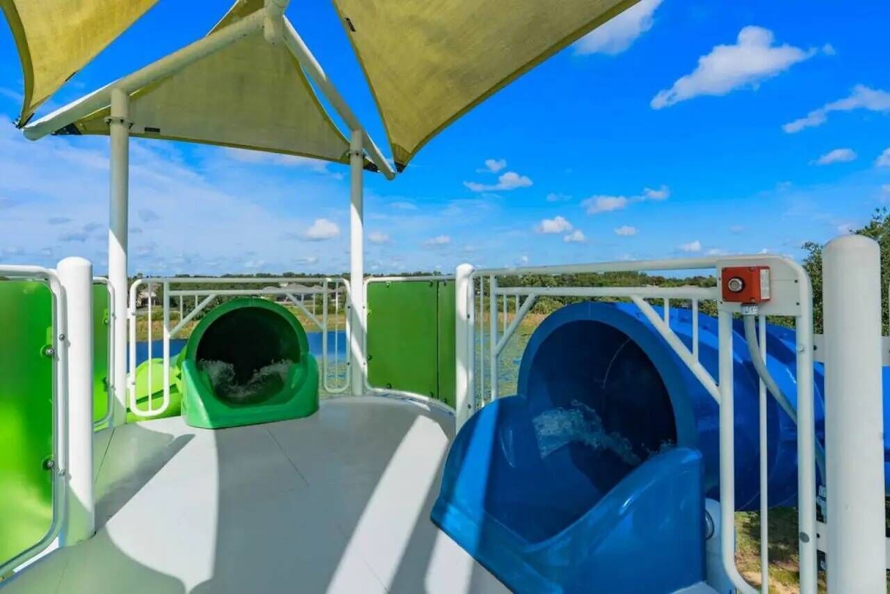 A water play area with slides and a green tube slide, surrounded by safety railings and a clear sky.