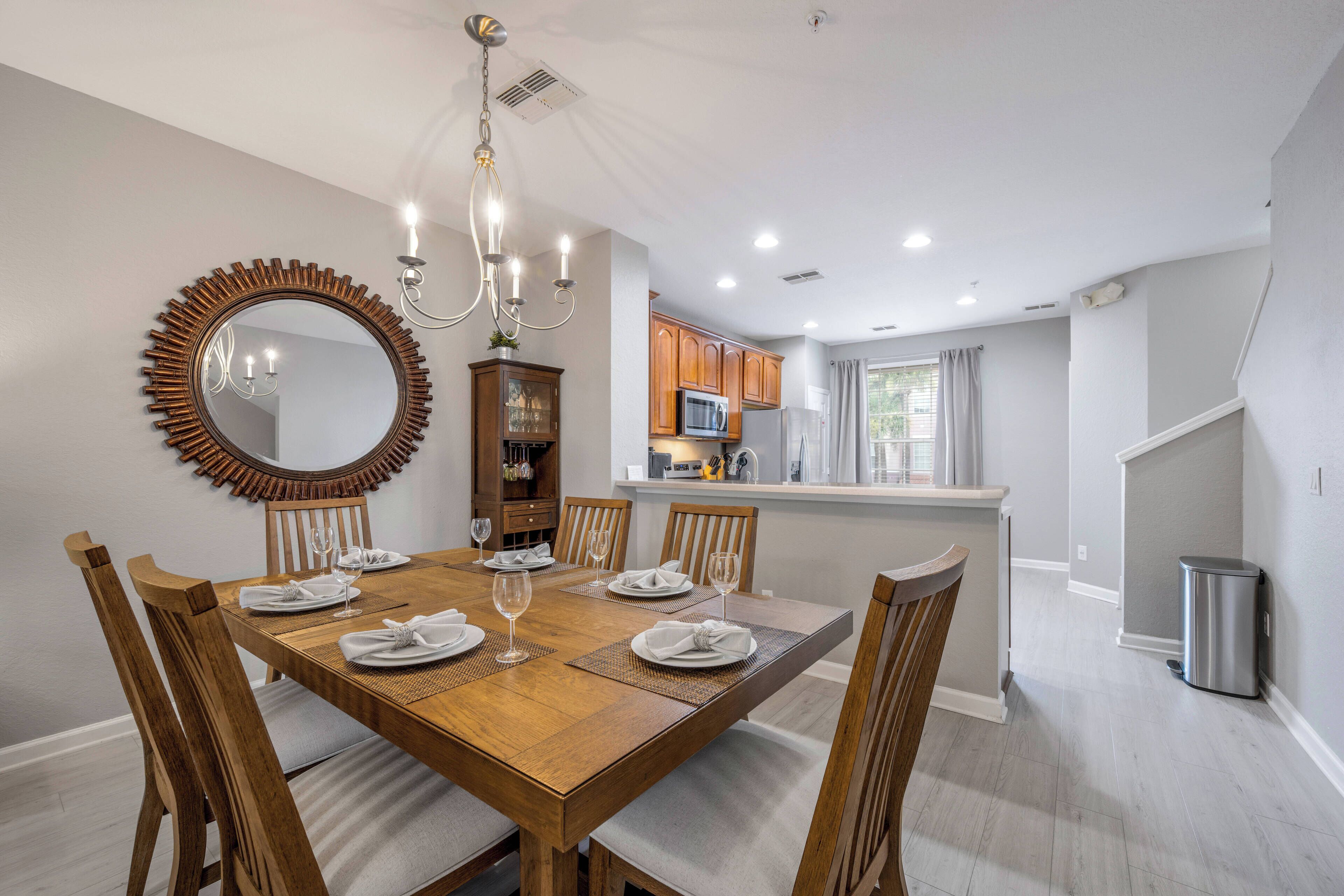 Un coin repas avec une table en bois dressée pour six personnes, entourée de chaises en bois. Un grand miroir avec un cadre décoratif est accroché au mur.