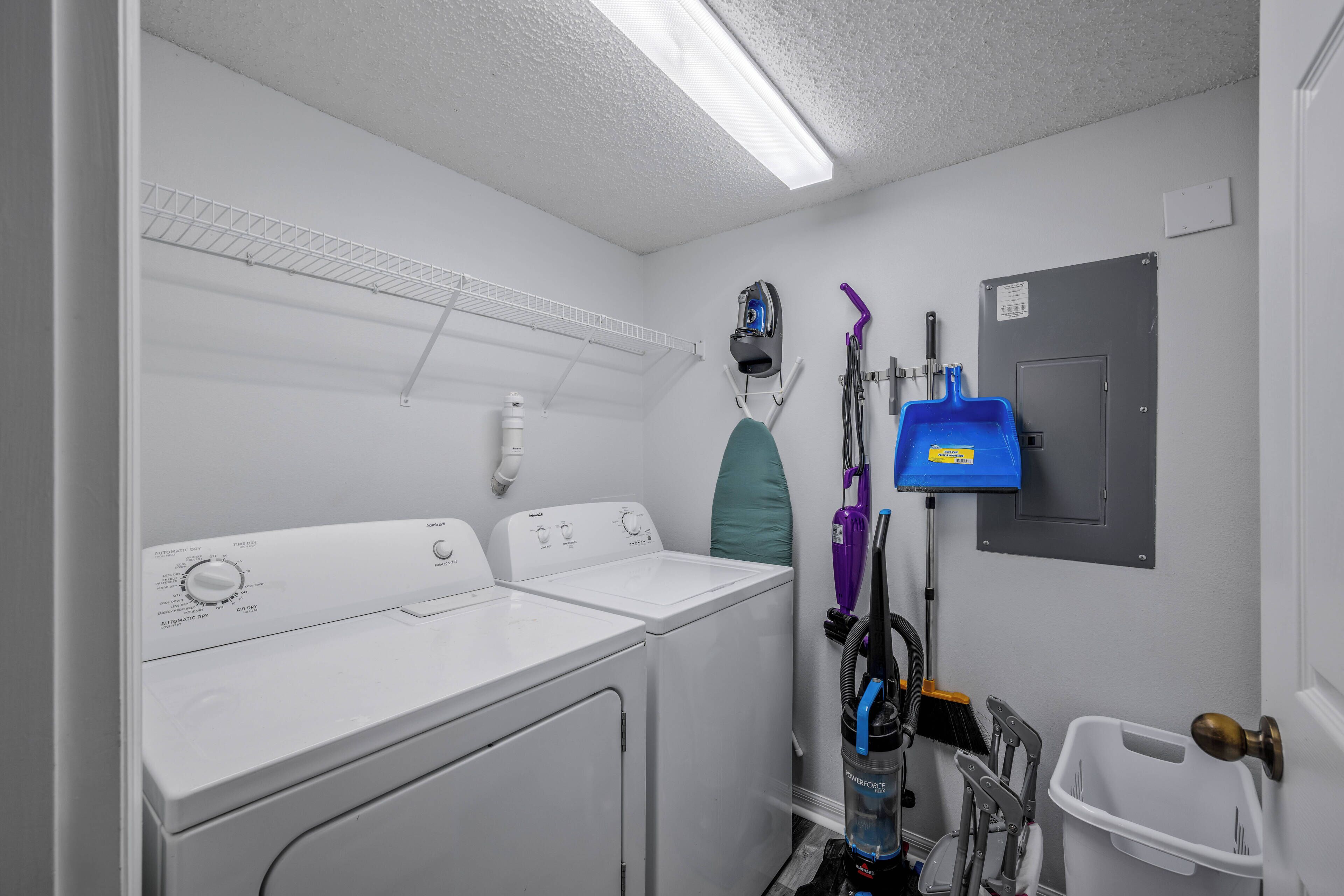 A laundry room with two white washing machines, a drying rack, a blue vacuum cleaner, and a white trash bin.