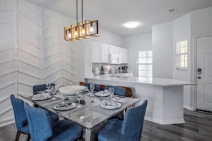 A modern kitchen with white cabinets, a central island, and a dining area with a glass table and blue chairs. - Ei8555 - 3BR House Near Disney (Kissimmee)
