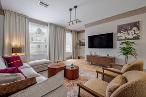 Una sala moderna con un sofá, una mesa de centro y sillones. Hay un televisor montado en la pared y una planta al lado.