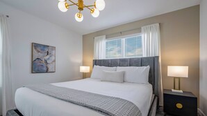 Een netjes opgemaakte slaapkamer met een groot bed, bedlampjes, een nachtkastje, een schilderij en een raam met gordijnen.