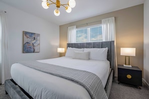 A neatly arranged bedroom with a large bed, bedside lamps, a nightstand, a painting, and a window with curtains. - Sl4359 - 5BR House With Pool Near Disney (Kissimmee)