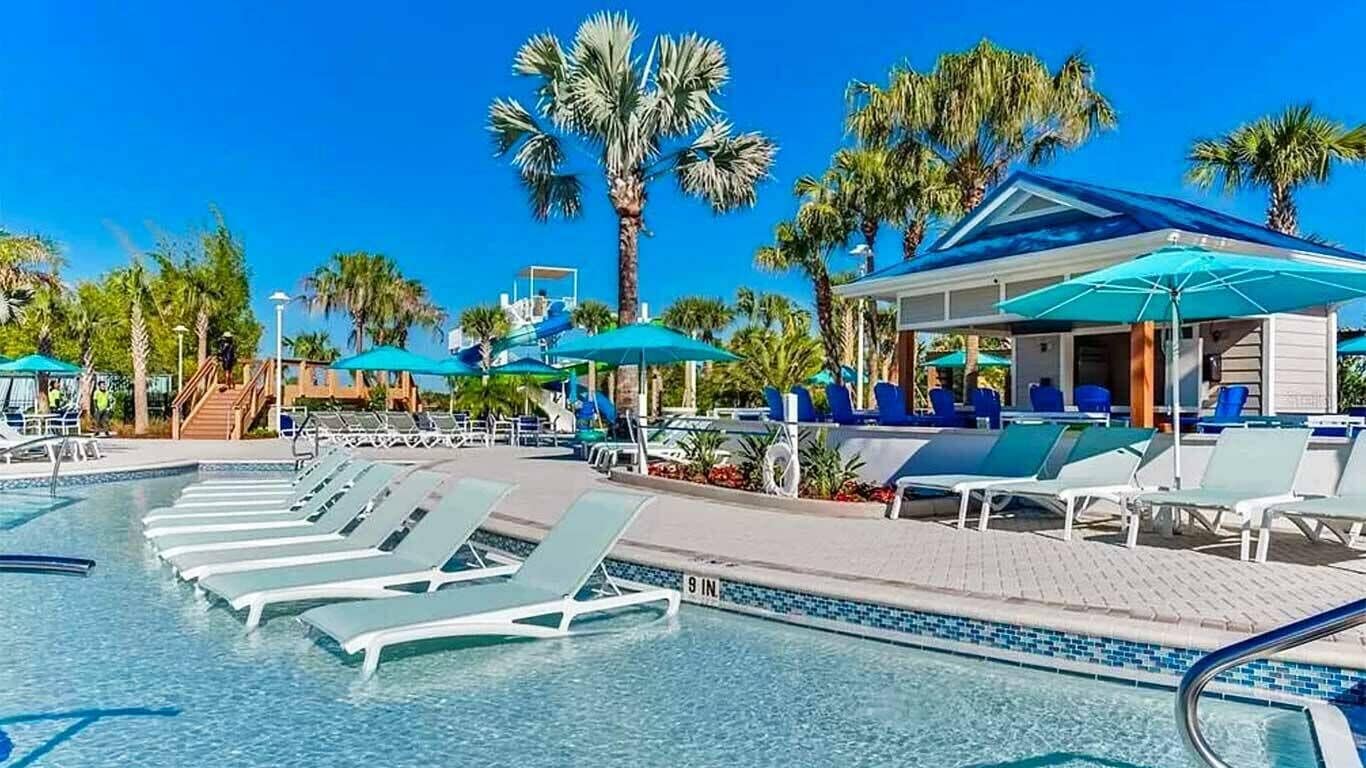 Un área de piscina con sillas de descanso y sombrillas, palmeras y un edificio con techo azul.