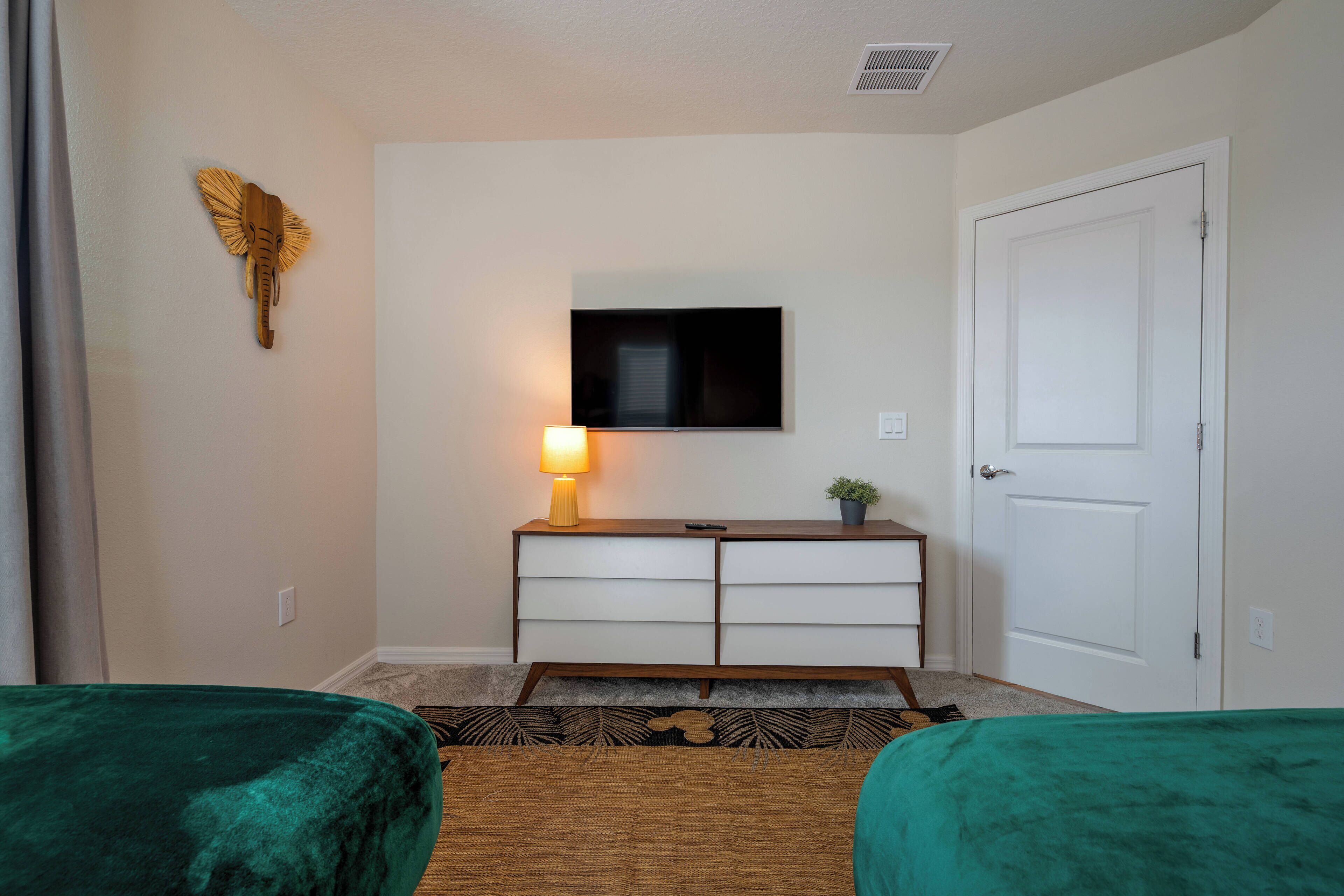 A hotel room with two green beds, a TV on the wall, a small table with a lamp, and a door.
