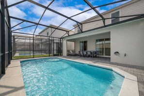 A swimming pool with a covered patio area, a table with chairs, and a view of a house in the background. - Wi3277 - 5BR House Near Disney With Private Pool (Davenport)