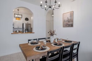 A dining area with a wooden table set for four, surrounded by black chairs. There is a chandelier above the table, a vase with dried flowers on the table, and a framed picture on the wall. - Wp304 - 2BR Fully Renovated Condo - Windsor Palms (Kissimmee)