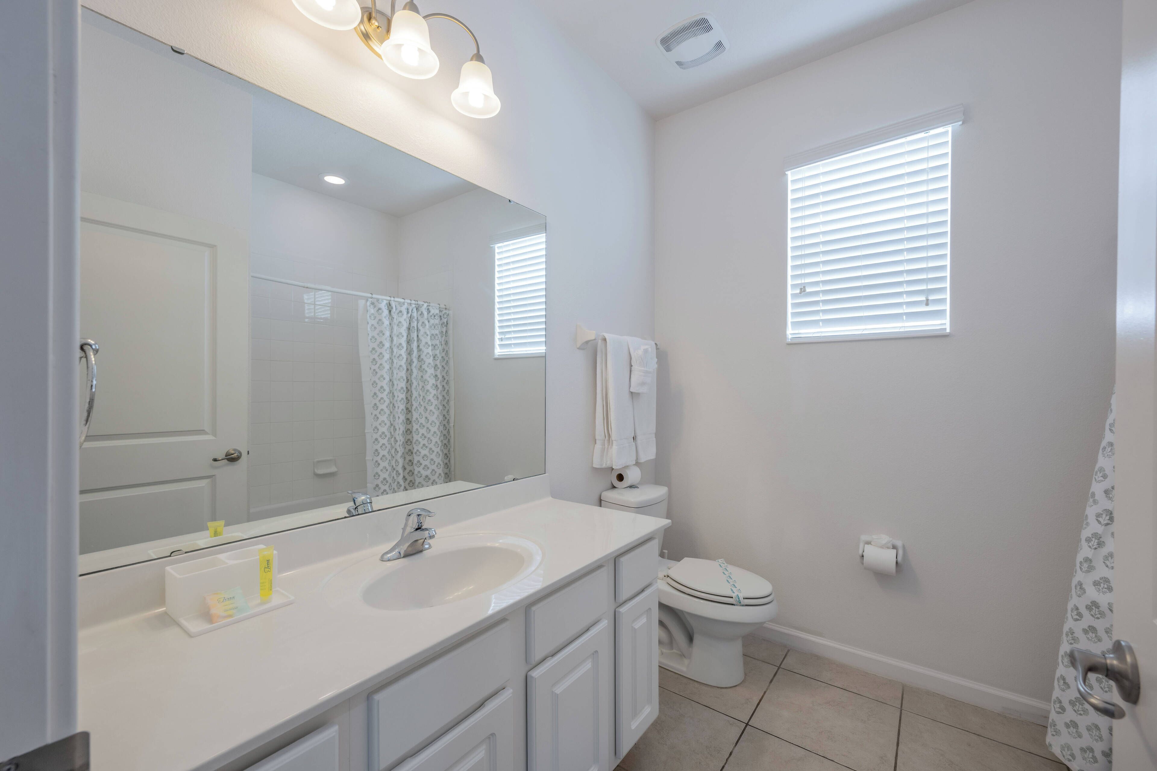 A modern bathroom with a white sink, mirror, and toilet. There are two windows with blinds, a shower curtain, and a towel rack.