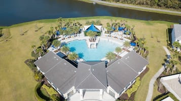An aerial view of a resort with a large central pool, surrounded by palm trees and a lake.