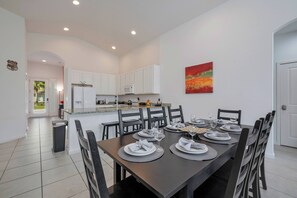 Un espace repas moderne avec une longue table dressée pour six personnes, des meubles blancs, un four à micro-ondes et un tableau abstrait rouge accroché au mur.