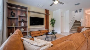 Una sala de estar con un sofá de cuero marrón, un televisor, un ventilador de techo y una escalera.