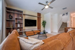 Ein Wohnzimmer mit braunem Ledersofa, einem Fernseher, einem Deckenventilator und einer Treppe.