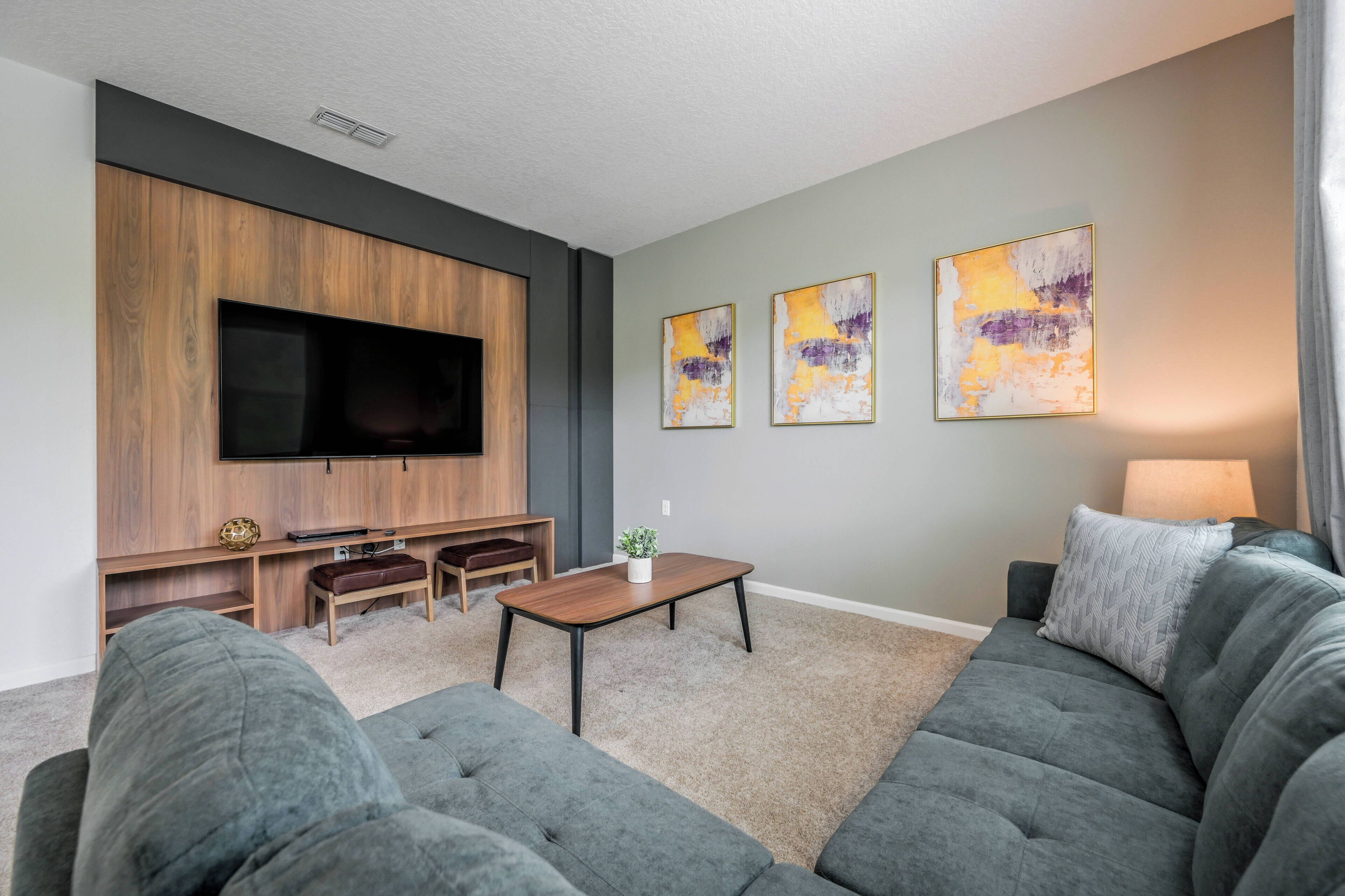 A modern living room with a large flat-screen TV, grey sofas, a wooden entertainment unit, and abstract wall art.