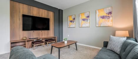A modern living room with a large flat-screen TV, grey sofas, a wooden entertainment unit, and abstract wall art.