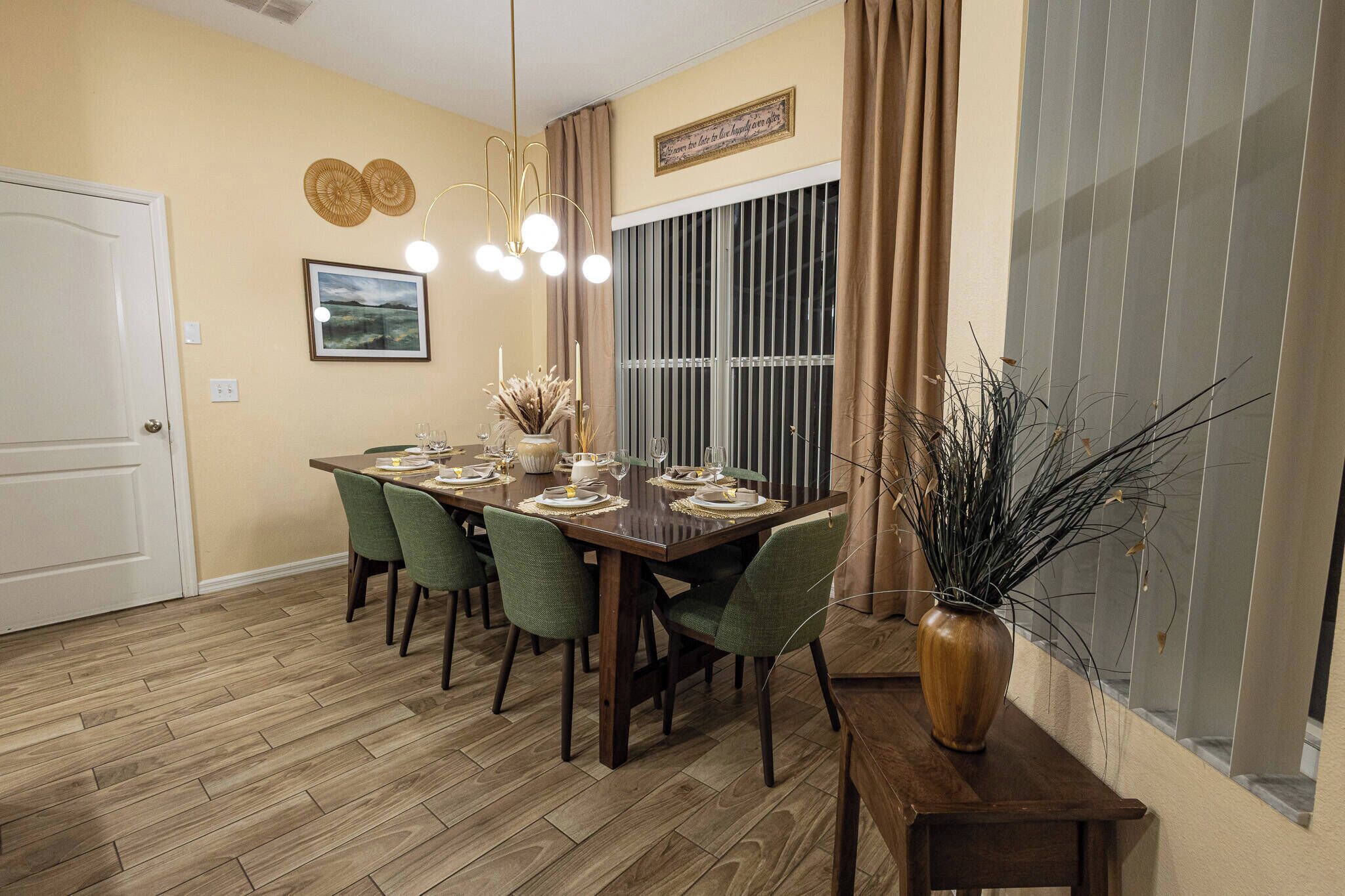 A dining area with a wooden table set for six, green chairs, a vase with dried plants, and a large window with vertical blinds.
