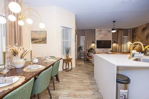 Un espace de repas moderne avec une table en bois dressée pour le dîner, une cuisine dotée d’un îlot central et un salon avec un canapé et un téléviseur.