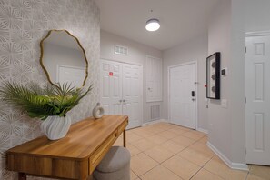 A hallway with a wooden console table, a white vase with a plant, and a mirror with a gold frame. There are white doors and a white wall with a geometric pattern. - Vc9-410 - 3BR Condo Near all Attractions (Orlando)