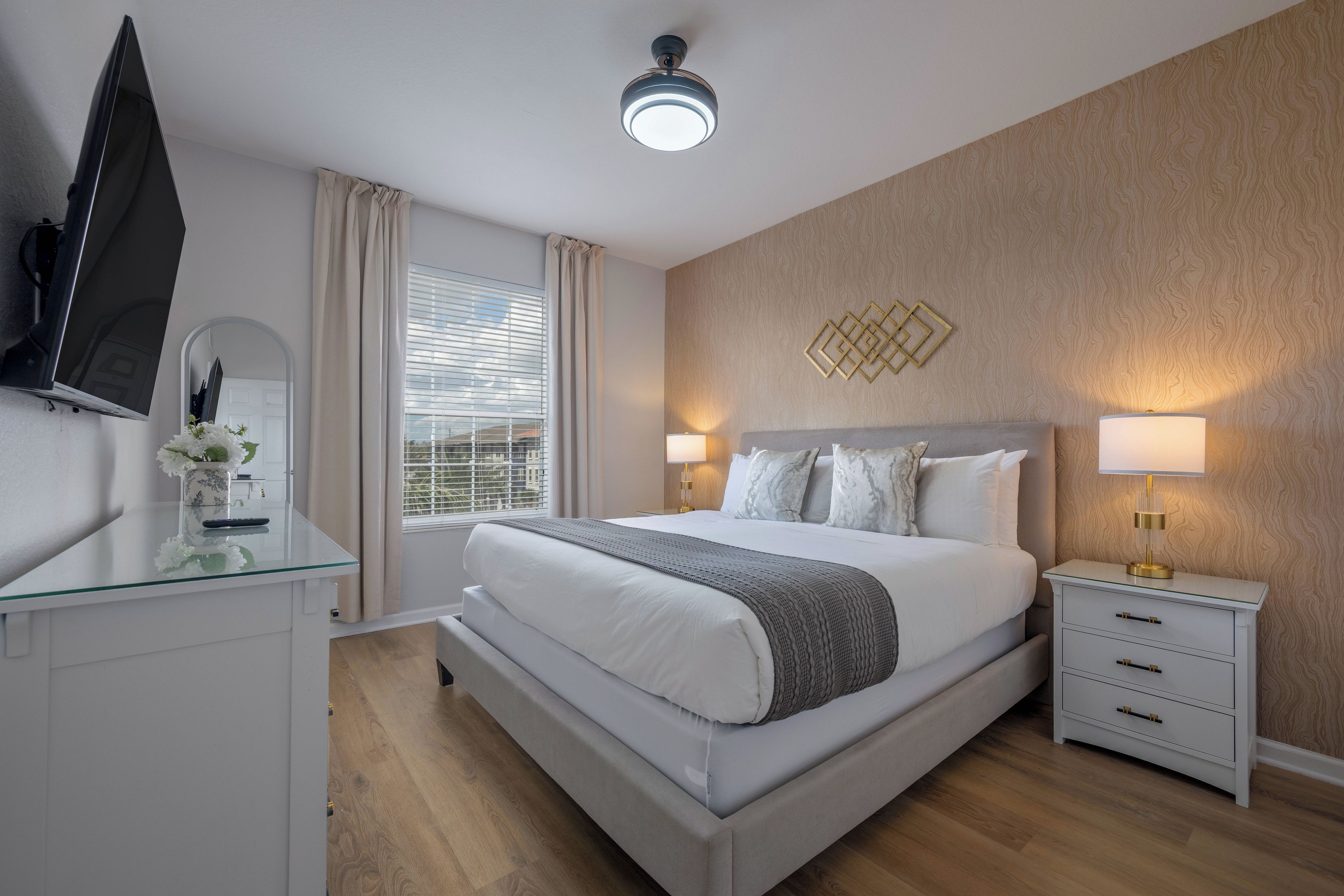 A modern hotel room with a large bed, bedside lamps, a flat-screen TV, and a glass desk with a vase of flowers.