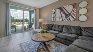 A living room with a grey sofa, a round coffee table with a plant, a ceiling fan, and a large window with a view of a park. - Sl4751-208 - 3BR Apartment Near Disney (Kissimmee)