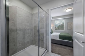 Ein modernes Badezimmer mit Duschkabine aus Glas, begehbarer Dusche und einem sichtbaren Schlafzimmer mit Bett und Fenster.