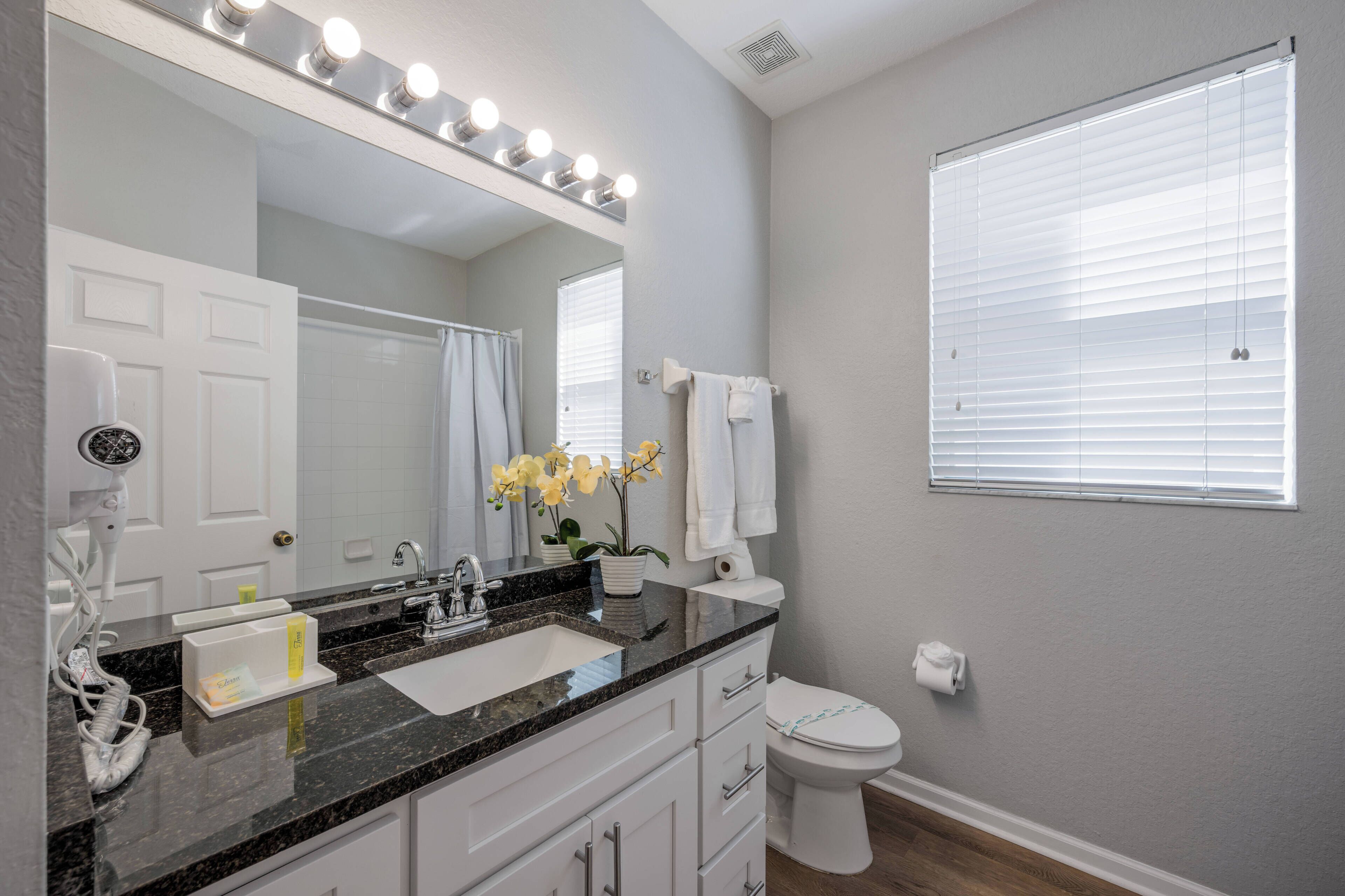 Une salle de bain équipée d’un lavabo, d’un miroir et de toilettes. Il y a des lampes au-dessus du miroir et une fenêtre avec des stores.
