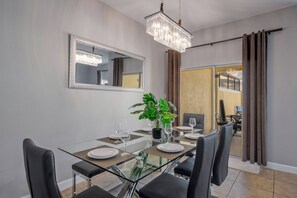 Ein Essbereich mit einem Glastisch, schwarzen Stühlen, einer Kristallleuchter, einem Spiegel und einer Pflanze.