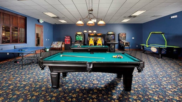 Une table de billard entourée de jeux d’arcade, une table de ping-pong et une table de baby-foot dans une pièce dont le sol est recouvert d’une moquette à motifs et les murs sont bleus.