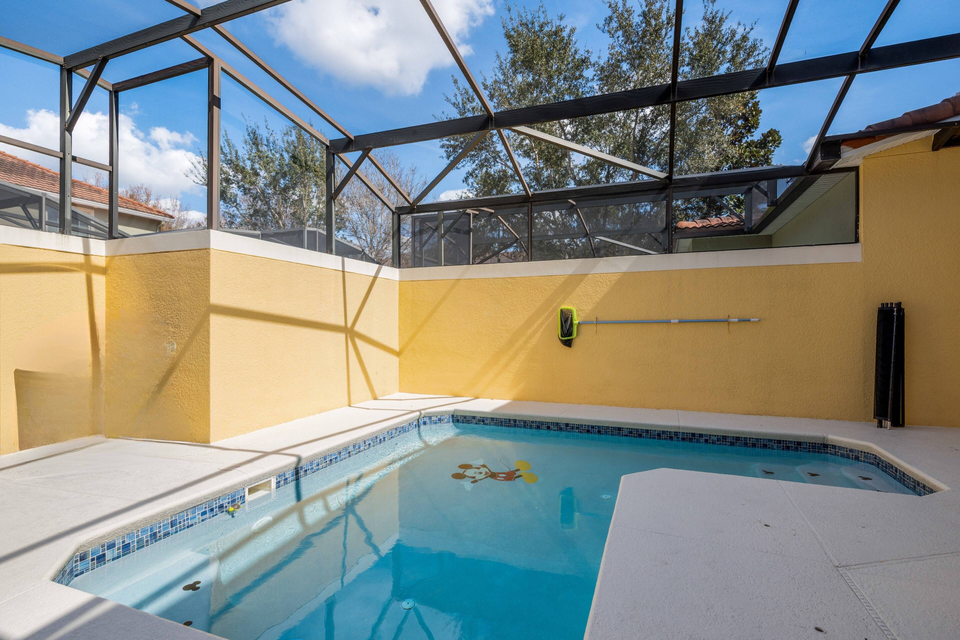A swimming pool with a glass ceiling, surrounded by a yellow wall, and a clear view of the sky and trees.