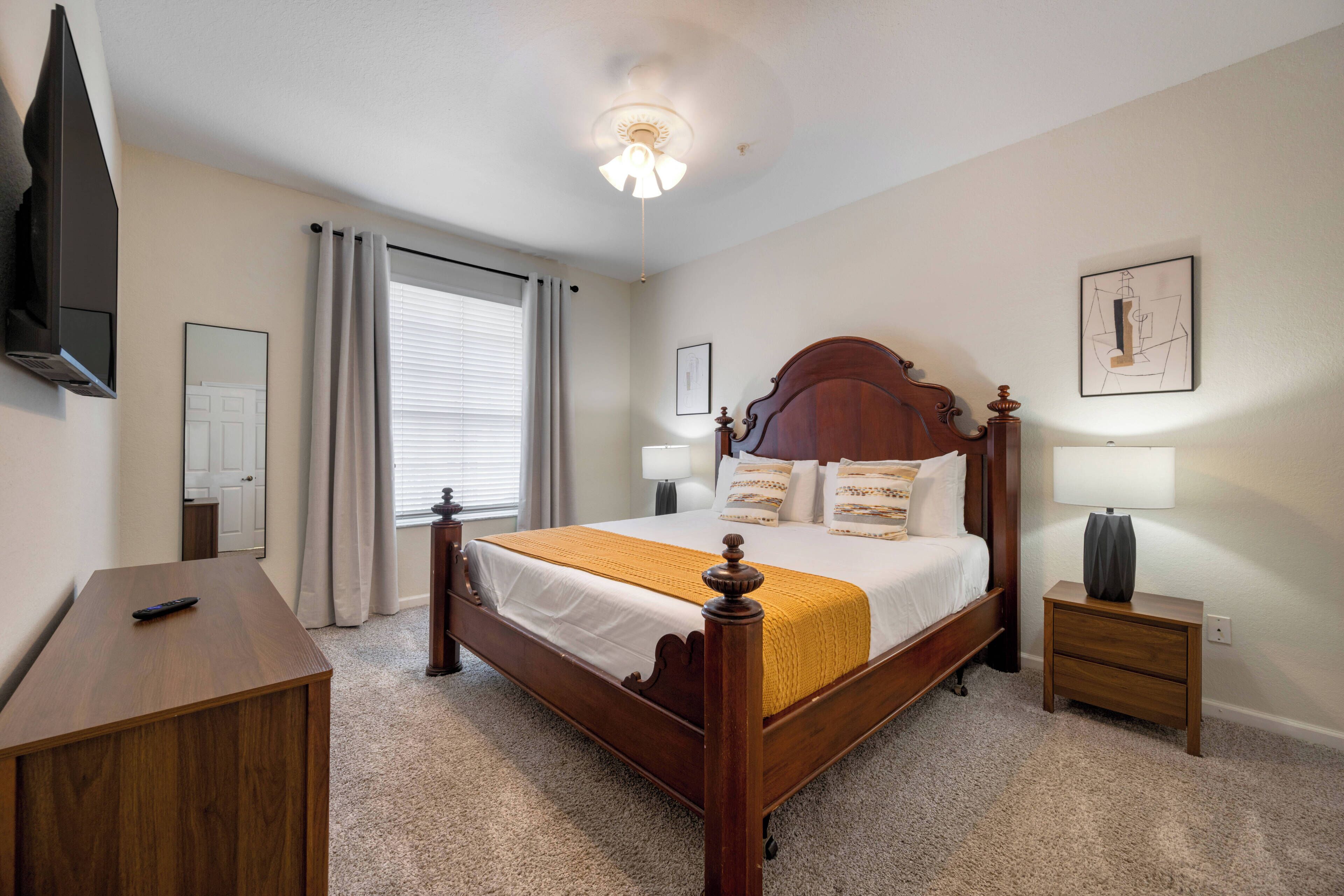 A bedroom with a wooden bed, a bedside table, a lamp, a TV mounted on the wall, and a window with curtains.