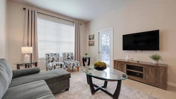 Una sala de estar con un sofá, dos sillones, una mesa de centro de vidrio, un televisor, un mueble de entretenimiento de madera y una ventana con cortinas.