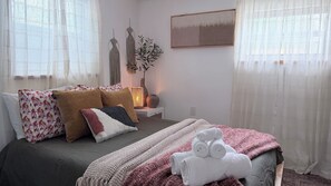 Een slaapkamer met een bed, kussens, een nachtkastje met een lamp en een raam met gordijnen.