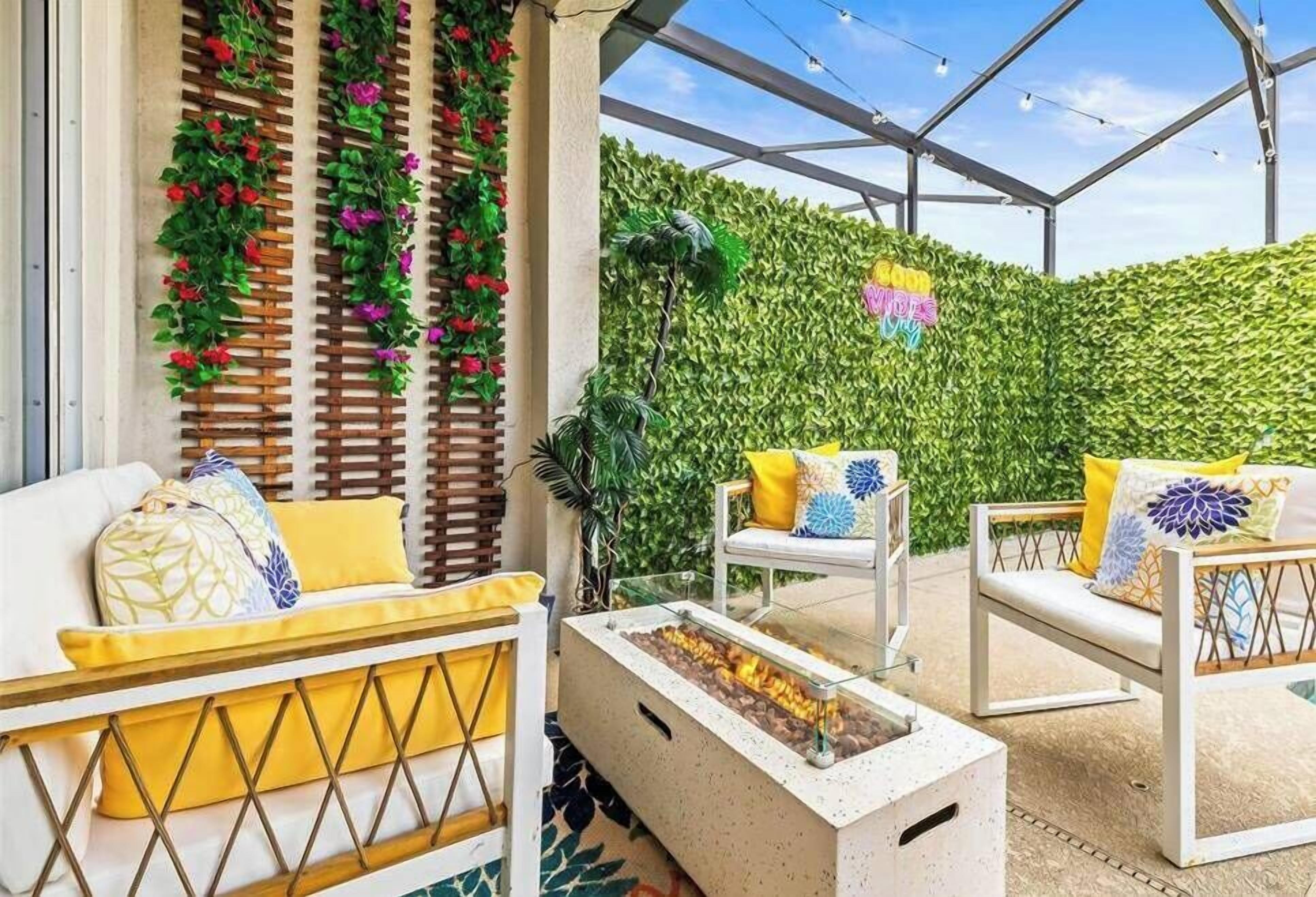 Un área de estar al aire libre con una chimenea, sillas blancas con cojines amarillos y una pared verde cubierta de plantas trepadoras.