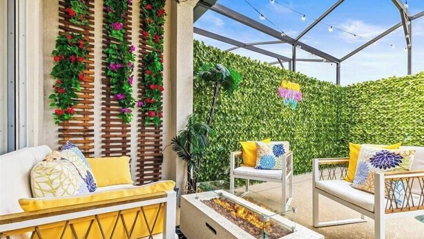 Un área de estar al aire libre con una chimenea, sillas blancas con cojines amarillos y una pared verde cubierta de plantas trepadoras.
