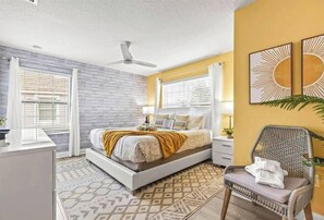 Un dormitorio con cama, mesitas de noche, una silla, ventilador de techo y una obra de arte en la pared.