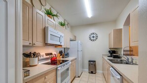 A modern kitchen with wooden cabinets, a microwave, stove, refrigerator, sink, and a trash bin. - Wp302 - 2BR Condo Near Walt Disney World (Kissimmee)