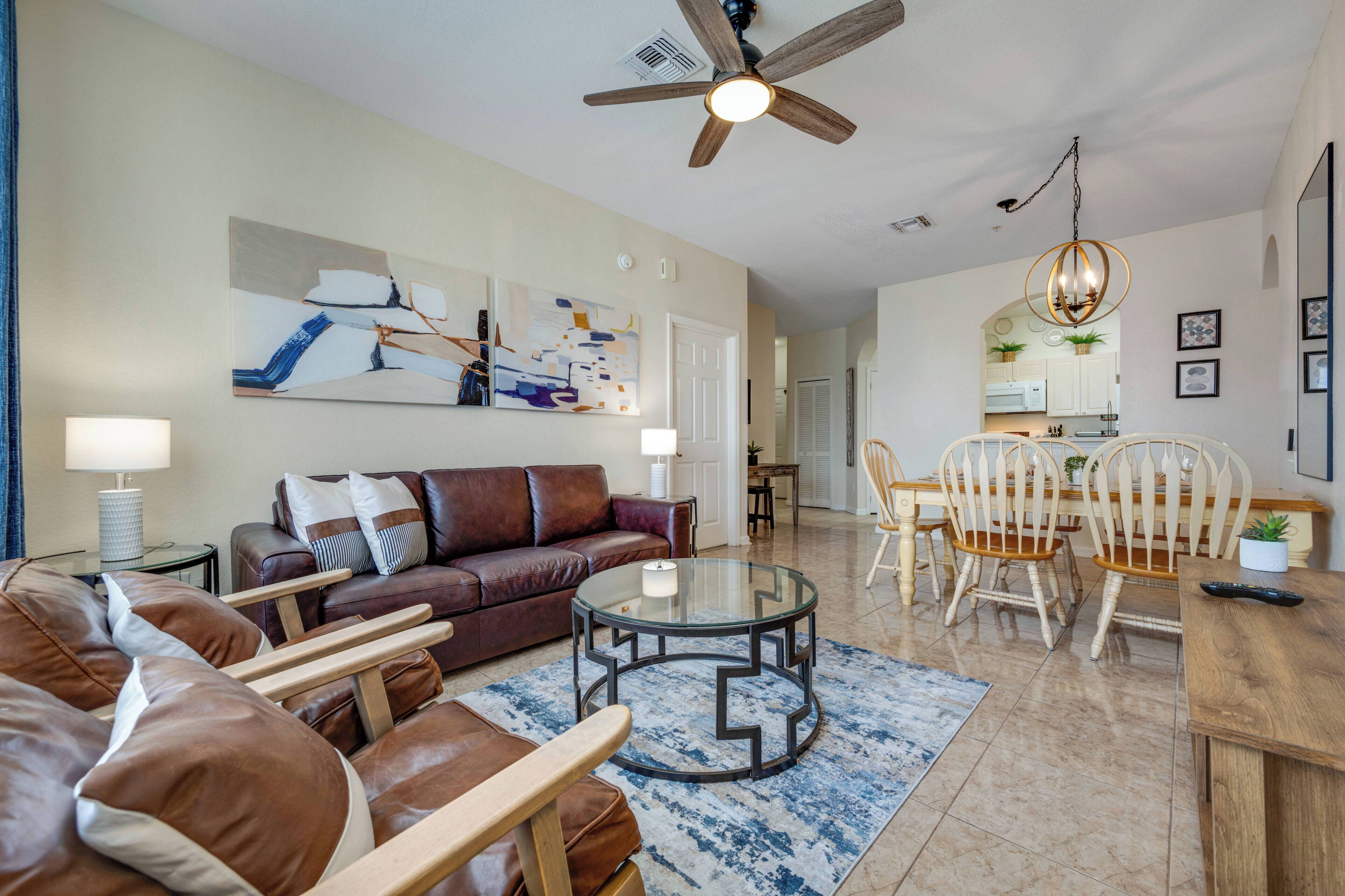 A living room with a brown leather sofa, a glass coffee table, and a dining area with wooden chairs and a chandelier.