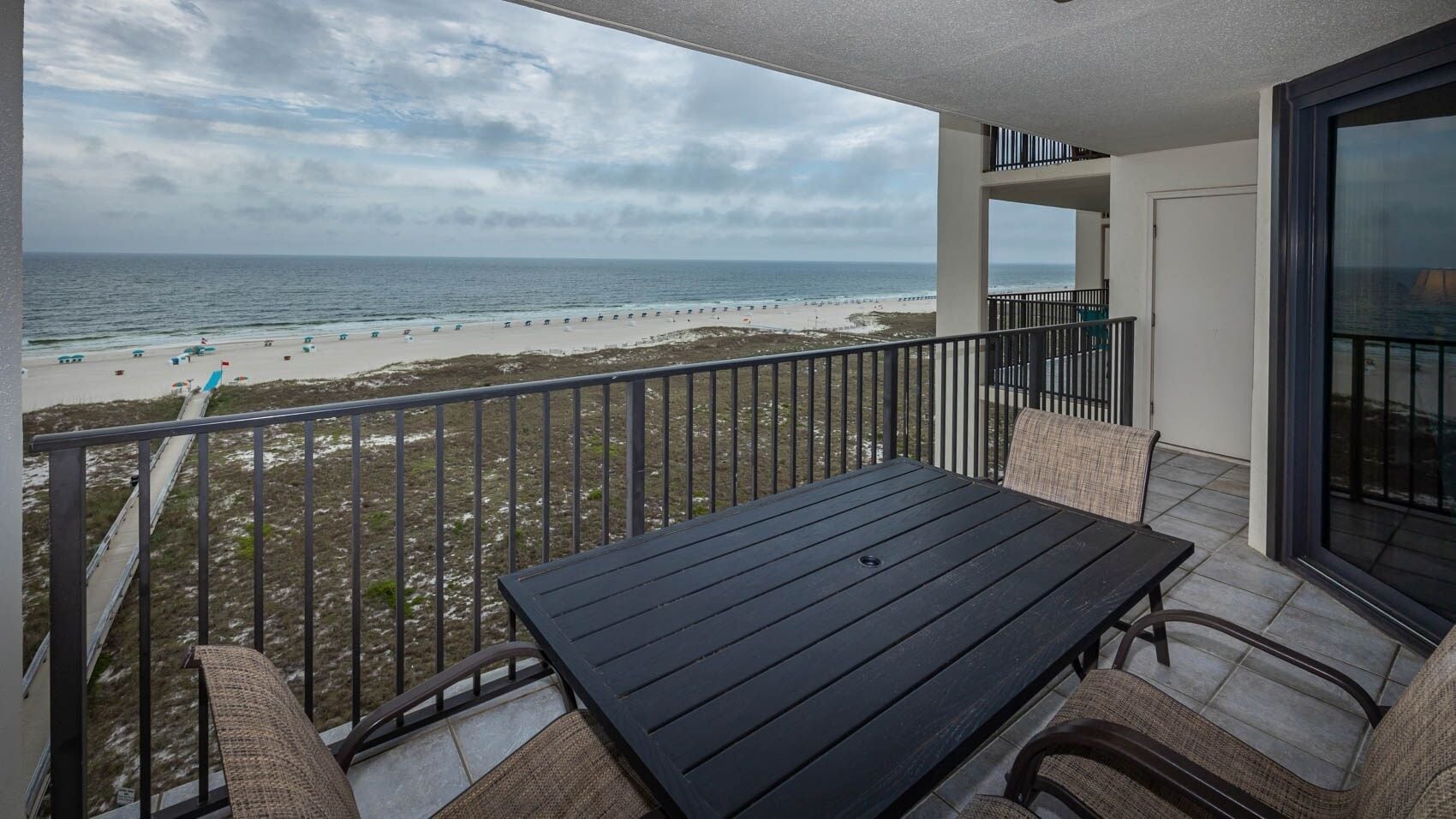 A balcony with a view of the beach, a table, and chairs.