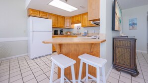 A kitchen with wooden cabinets, a white refrigerator, a central island with stools, and a decorative cabinet. - Phoenix Iii 3043 1 Bedroom Condo (Orange Beach)