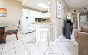 A modern kitchen with white cabinets, a granite countertop, and a built-in oven. There are two white bar stools and a dining table with a glass top. A flat-screen TV is mounted on the wall, and a refrigerator is visible in the background. - Phoenix Vii 71514 1 Bedroom Condo (Orange Beach)